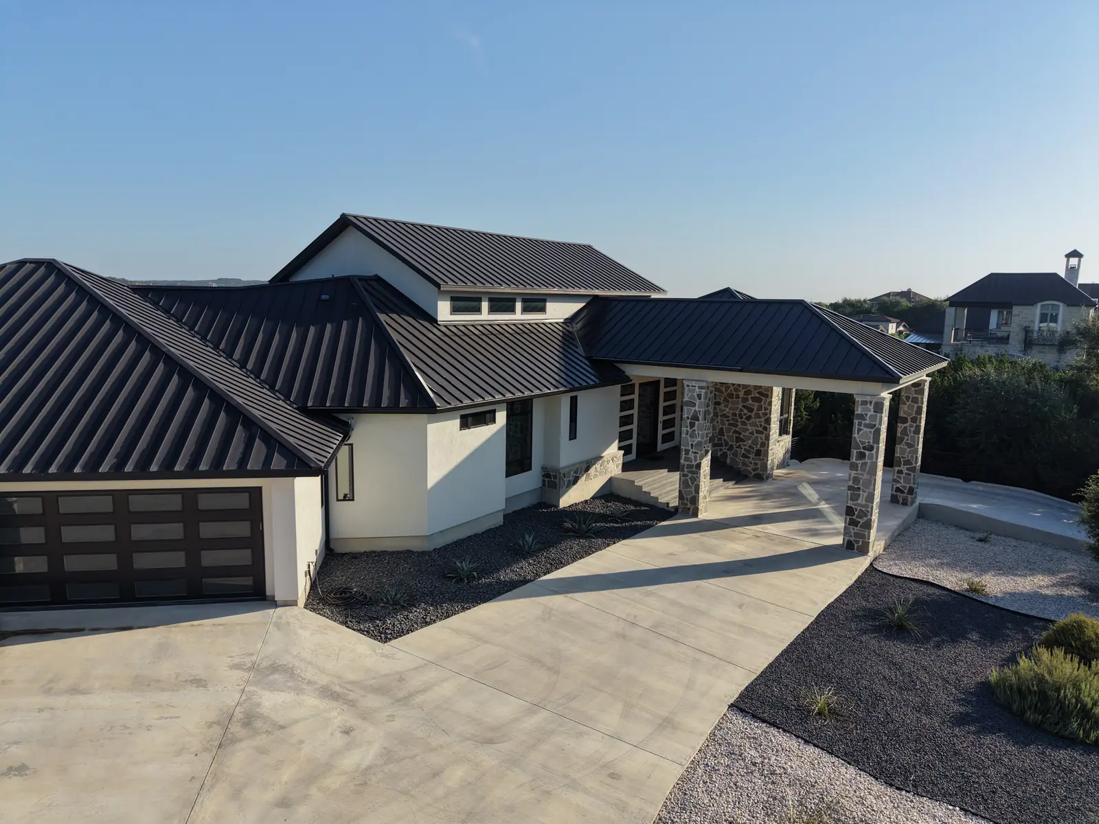 Front elevation of a completed modern custom home with black metal roof, stone accents and a wide concrete driveway
